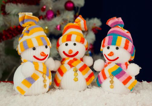 Three snowmen in front of the decorated white evergreen tree over the blue background