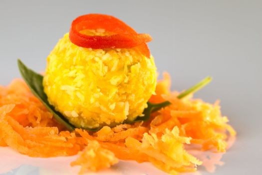 Pumpkin coconut ball garnished with a red pepper slice served on basil leaf and grated carrot (Selective Focus, Focus on the front of the pepper garnish and the pumpkin ball)