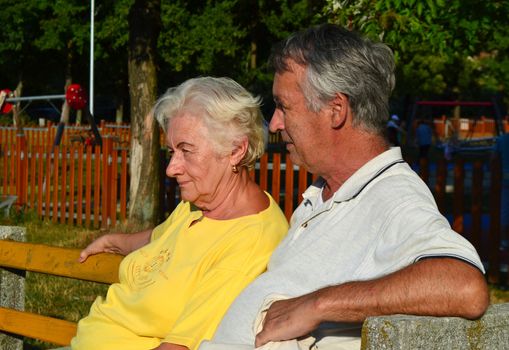 Elderly couple (60s) sitting and relaxing on a bench, enjoying the afternoon sunshine.