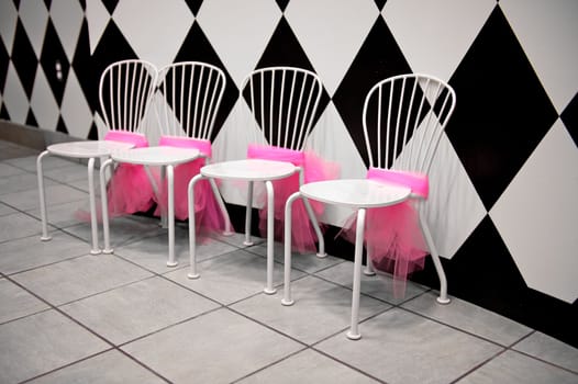 A row of four white chairs with pink bunting or ribbons tied around them next to a black and white wall