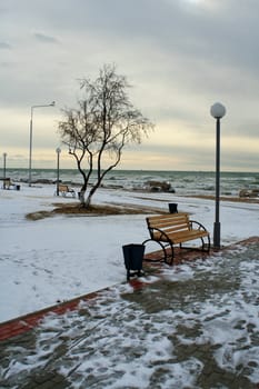 Deserted Bench On Winter Promenade with the cold ocean behind and snow on the ground.