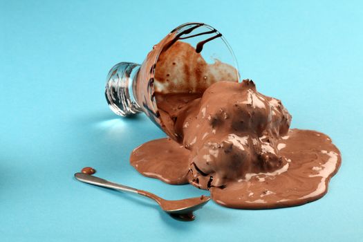 Photo of a bowl of melting chocolate ice cream tipped over and melting across a blue table.