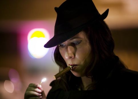 Woman in a fedora lighting a cigar with a match.