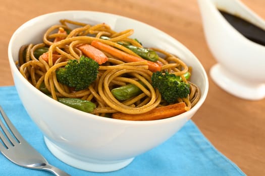 Vegetable and wholewheat spaghetti stir fry in white bowl with soy sauce in the back (Selective Focus, Focus on the broccoli floret on the left)