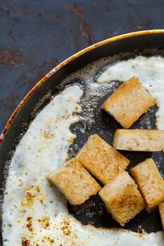 Frying pan full of scramble eggs from two eggs with a small pieces of toasted bread. Stands on grunge metal background.