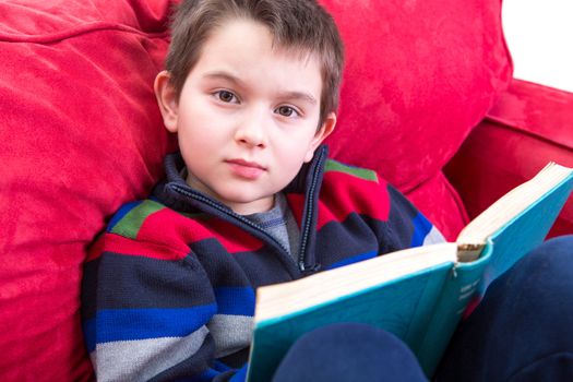 Kid looking at camera while reading a book on the red couch. He has a meaningful look on his face, perhaps distracted.