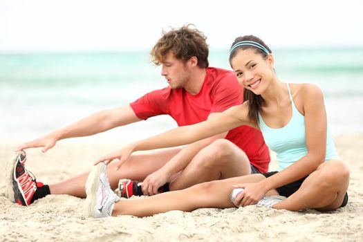 Couple training on beach stretching legs after running. Young man and woman during summer workout. Asian female fitness model, Caucasian male fitness model.