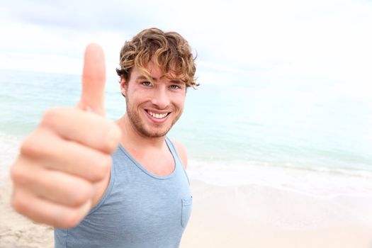 Young man thumbs up. Smiling happy sporty man giving thumbs up success sign to camera during training outside on beach. Handsome male fitness model in his 20s.