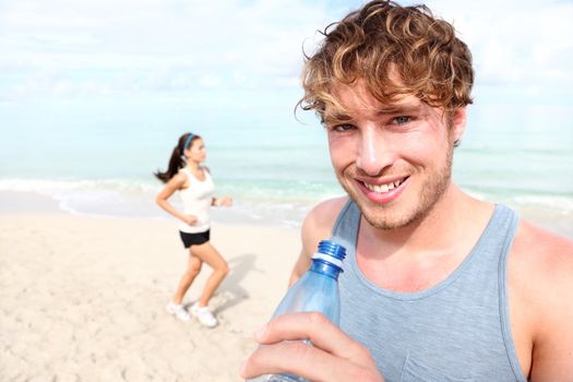 Running couple. Man drinking water smiling happy, woman runner in background. Young male fitness model in his 20s outdoors on beach