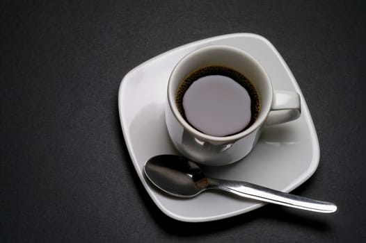 White coffee cup with saucer and spoon on dark grey background closeup