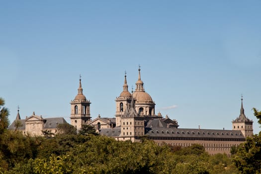 San Lorenzo de El Escorial Monastery from Casita del Infante. The towers of the church and monastery are set of by a bright blue sky.