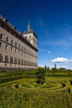 San Lorenzo de El Escorial Monastery with its formal gardens. The towers of the monastery are set of by a bright blue sky with a few white clouds.