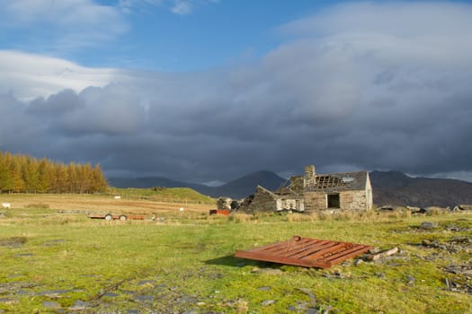 A rusty gate lays on green grass in front of  a derelict quarry building with dark mountains and a moody sky in the background.