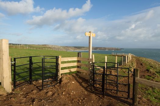 A coast footpath with two gates and a dual directional sign with green fields leading down to a sea bay backed by a blue cloudy sky.