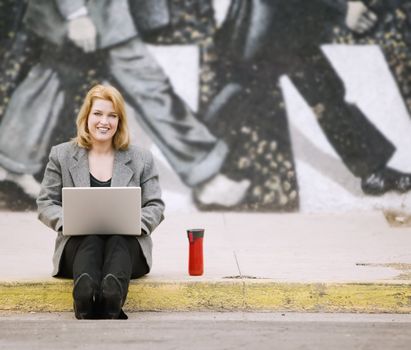 Pretty smiling woman with a laptop computer sitting on the sidewalk