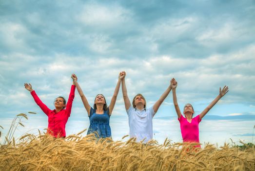 Four young people staying with raised hands at a wheat field at sunset time