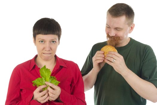 Bearded happy man with a hamburger smiles at a sad woman with lettuce leaves in her hands