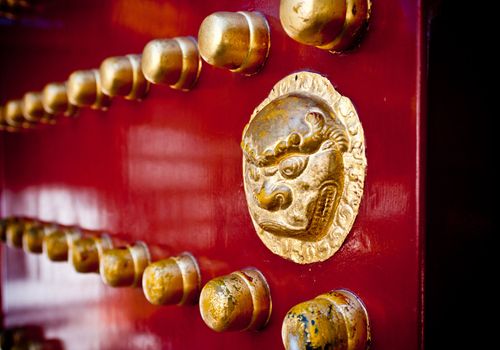 A brass-painted lion door handle on a red gate in an Oriental palace.
