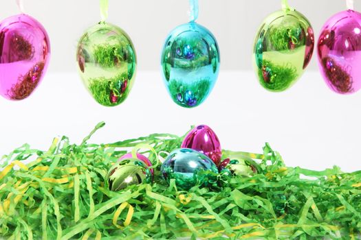 Colourful shiny metallic Easter Eggs hanging in a row above additional eggs nestling in a bed of green straw for a vibrant seasonal greeting card