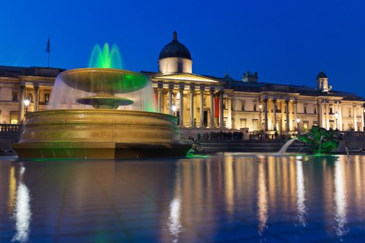 Trafalgar Square in the evening, the National Gallery. In the foreground - illuminated fountain and pool, reflecting the sky and lights. London, UK