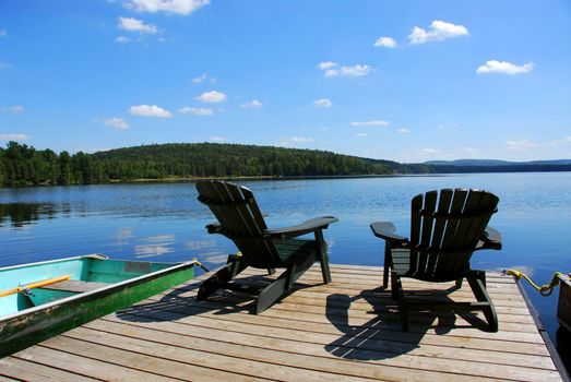 Two adirondack wooden chairs on dock facing a blue lake with clouds reflections