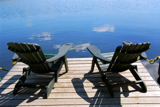 Two adirondack wooden chairs on dock facing a blue lake with clouds reflections