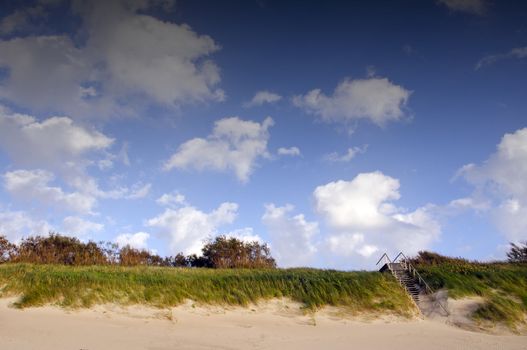 Coastal dunes and stairs leading up covered with sea sand. Blue sky with clouds.