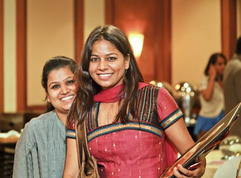 Two smiling Indian pretty hostesses at an event with clip board and mobile cell phone ushering guests, crop margin and empty copy space