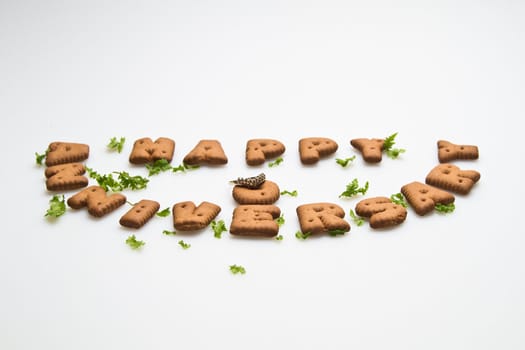 Angled view of Happy anniversary wording by brown biscuits, ring and green leaves on white surface