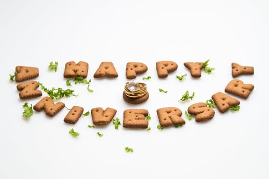 Angled view of Happy anniversary wording by brown biscuits, diamond ring and green leaves on white surface