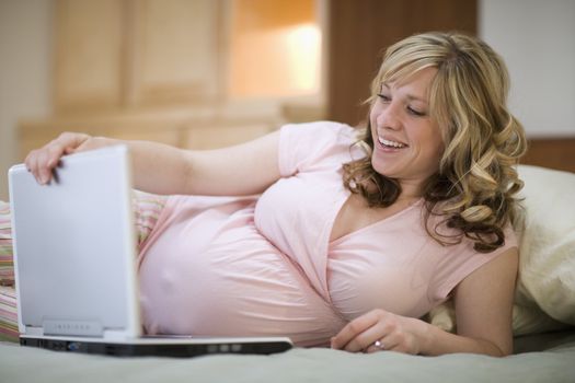 Smiling pregnant woman resting on bed and using laptop