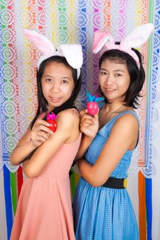 Cute Asian bunny girls hold Easter eggs in front of colorful flower shaped backdrop.