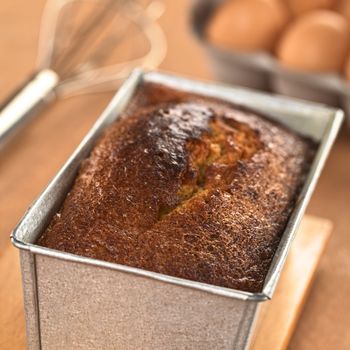 Freshly baked pound cake in baking pan with eggs and beater in the back (Selective Focus, Focus one third onto the surface of the cake) 