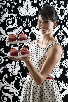 young woman is stylist 60's inspired clothing, serving a plate of velvet cupcake
