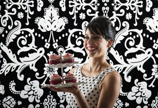 young woman is stylist 60's inspired clothing, with a big smile, holding a plate of velvet cupcake