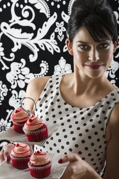 young woman is stylist 60's inspired clothing, serving a two storie plate of velvet cupcake