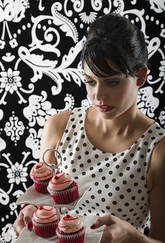 young woman is stylist 60's inspired clothing, concentration on not dropping a plate of velvet cupcake