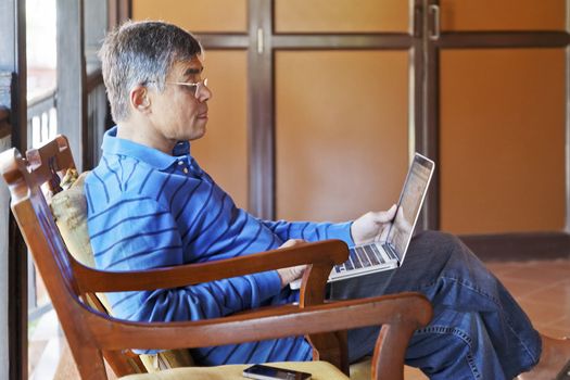 horizontal portrait of middle aged asian gentleman sat in captains chair on a a veranda with lap top compyter and mobile phone, copy space and crop margins