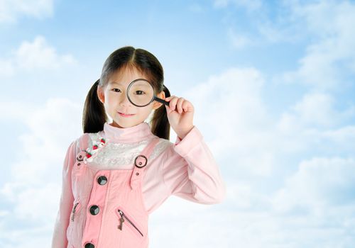 A little Asian girl peers at the camera through a magnifying glass in summer day, blue sky background