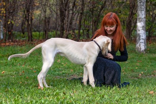 A girl in a black dress and white saliki pup in a forest 
