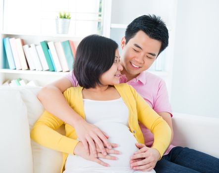 Mom and Dad with hands on the baby, sitting on sofa, Asian pregnant couple living lifestyle.