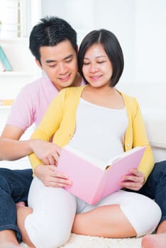 Asian pregnant couple reading book together, sitting on sofa at home.