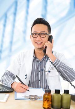 Young Southeast Asian medical doctor writing a note while calling on the phone