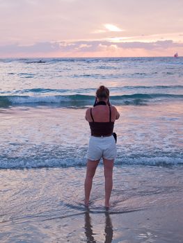 Attractive woman on a beach taking pictures with camera facing beautiful seascape sunset