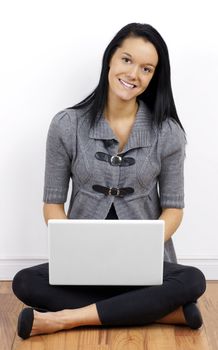 Friendly smiling beautiful young brunette woman or student looking at camera and sitting on wood floor with laptop against white wall.