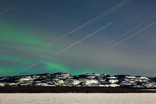 Astrophotography star trails with green glowing display of Northern Lights or Aurora borealis in moon lit night over boreal forest or taiga winter landscape of Lake Laberge  Yukon Territory  Canada