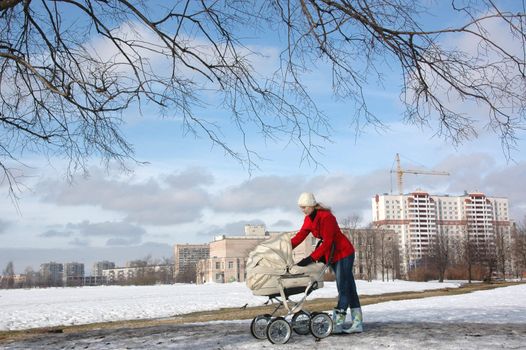 Wide shot of happy young mother in red coat with baby in buggy walking in winter park