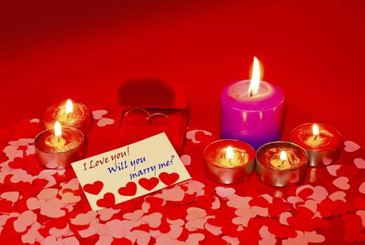 Two rings and a card with marriage proposal with six candles on the red background