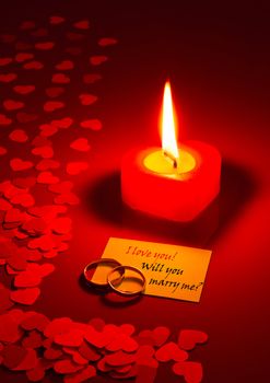 Two rings and a card with marriage proposal with a candle on the red background