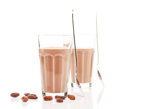 two glasses with chocolate milk and chocolate beans on white background with reflection